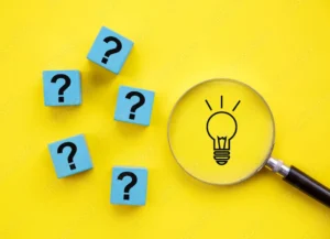 A bright yellow background with five light blue square blocks scattered on the left side, each displaying a large black question mark. On the right side, a clear magnifying glass with a black handle is positioned over the yellow surface, and the magnified area shows a simple black outline of a lightbulb with radiating lines above it, suggesting an idea or solution. The arrangement implies a search for answers or solutions to questions.