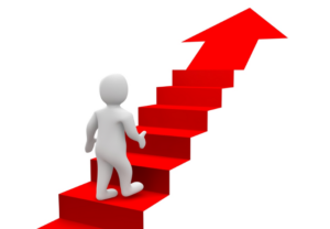 A 3D rendering on a white background depicts a small, featureless white figure with articulated limbs walking up a bright red staircase. The staircase begins at the bottom left and ascends diagonally towards the top right, where the final step seamlessly transitions into a large, solid red arrow pointing upwards and slightly to the right. The figure is positioned on the lower steps, appearing to be in the process of climbing towards the arrow, symbolizing progress and the achievement of goals.