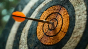 A close-up shot focuses on the center of a circular target made of weathered wood, showing concentric rings of black and a central orange bullseye. A brown arrow with a vibrant orange fletching is lodged firmly in the bullseye, its tip embedded in the wood. The arrow appears to have been in motion at the time of capture, resulting in a slight blur of the orange fletching. The background is softly blurred, suggesting an outdoor setting with greenery out of focus.