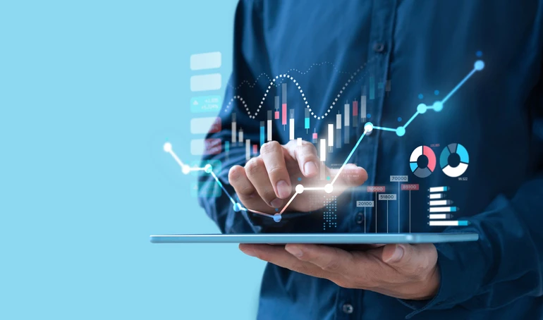 A person wearing a blue button-down shirt holds a tablet horizontally, with their right hand touching a glowing point on a holographic line graph that is projected above the tablet's screen. The holographic display includes various financial data visualizations: a dynamic line graph with upward trending arrows and percentage changes, a candlestick chart with red and green bars, a wavy white line, and two circular charts resembling pie charts with different colored segments. Below the line graph are translucent bar charts with numerical values. The background is a solid light blue.